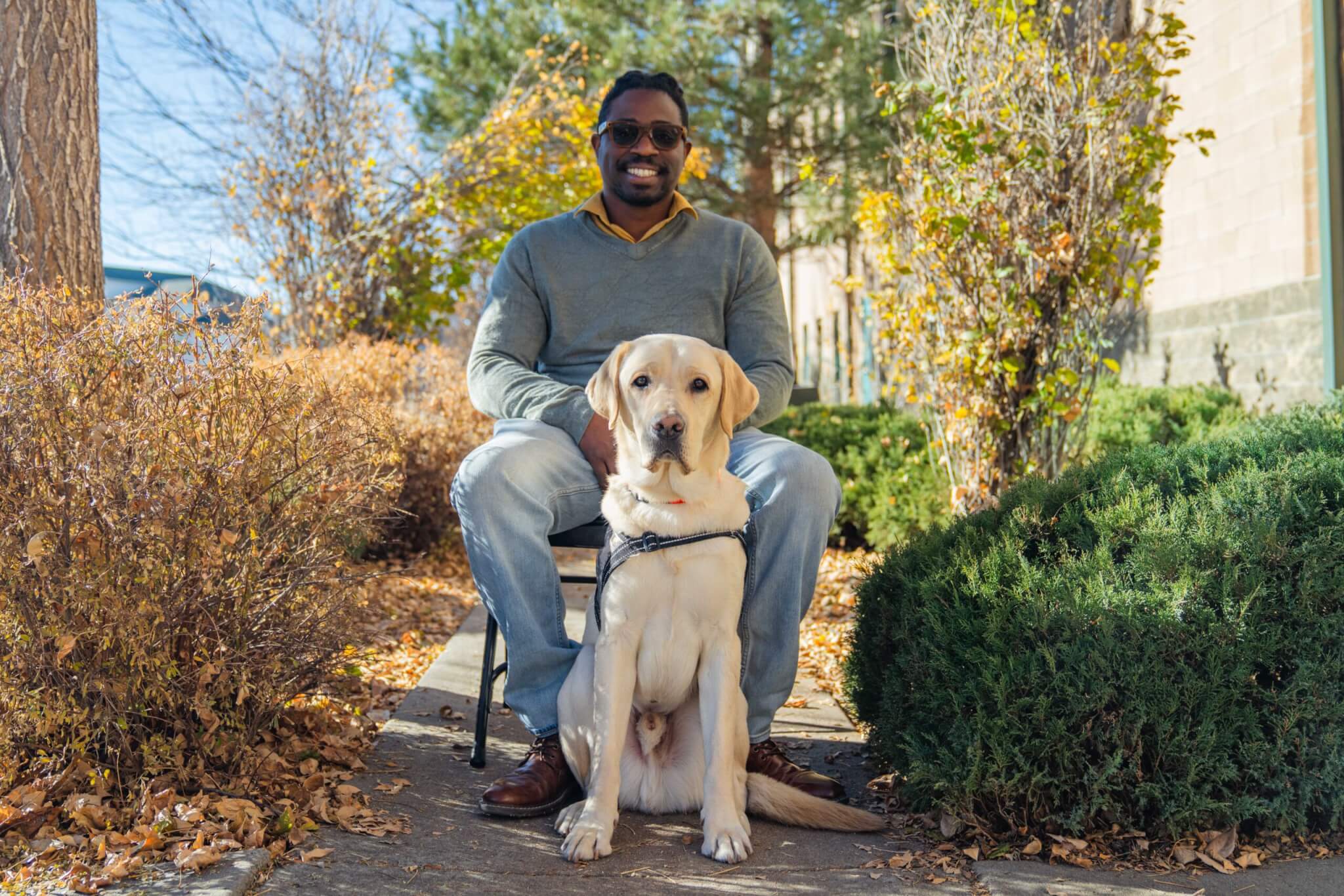 U.S. Army veteran man sitting on a chair between plants. Sitting in front of him is a male yellow Labrador Retriever wearing a Freedom Service Dogs vest.
