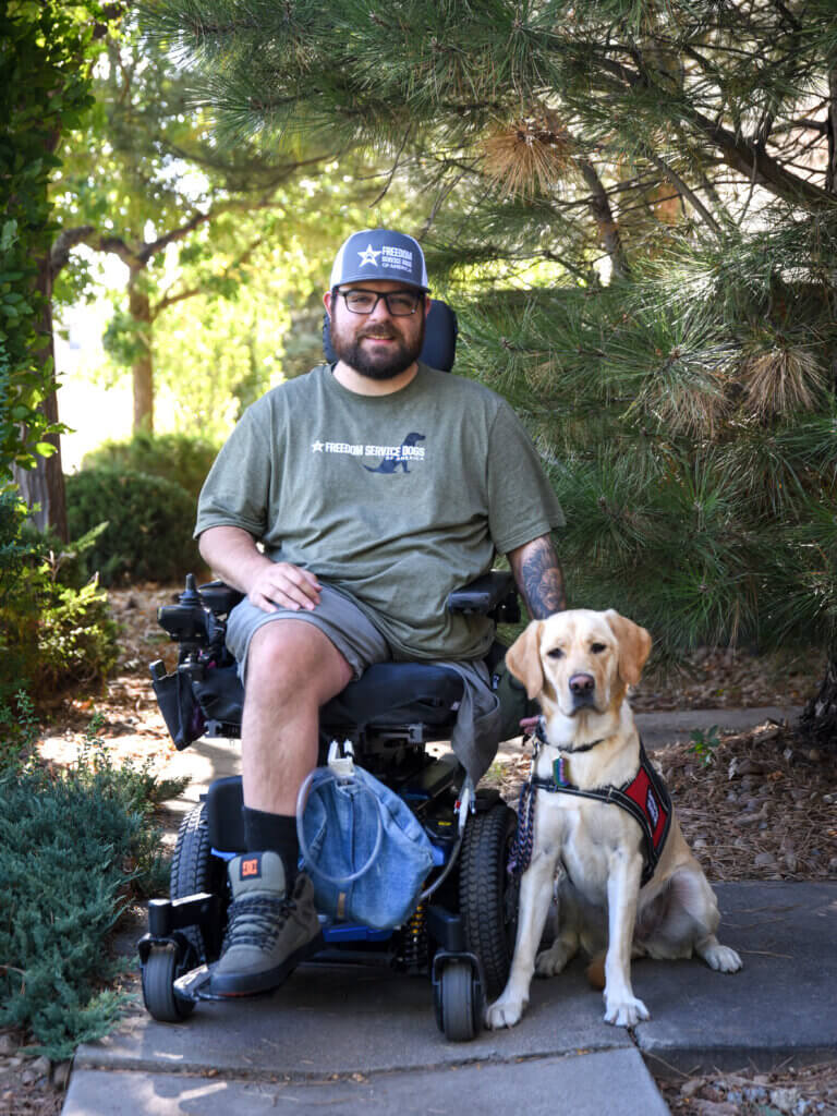 Yellow Labrador Retriever Freedom Service Dog sitting beside a man in a power chair wearing Freedom Service Dogs gear.