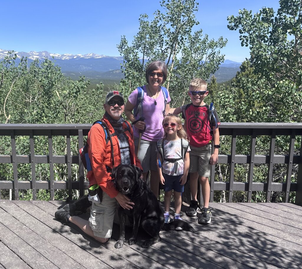 A man, woman, and two kids stand on a patio with a Colorado mountain background with a black and tan Labrador Retriever Skilled Companion Dog.