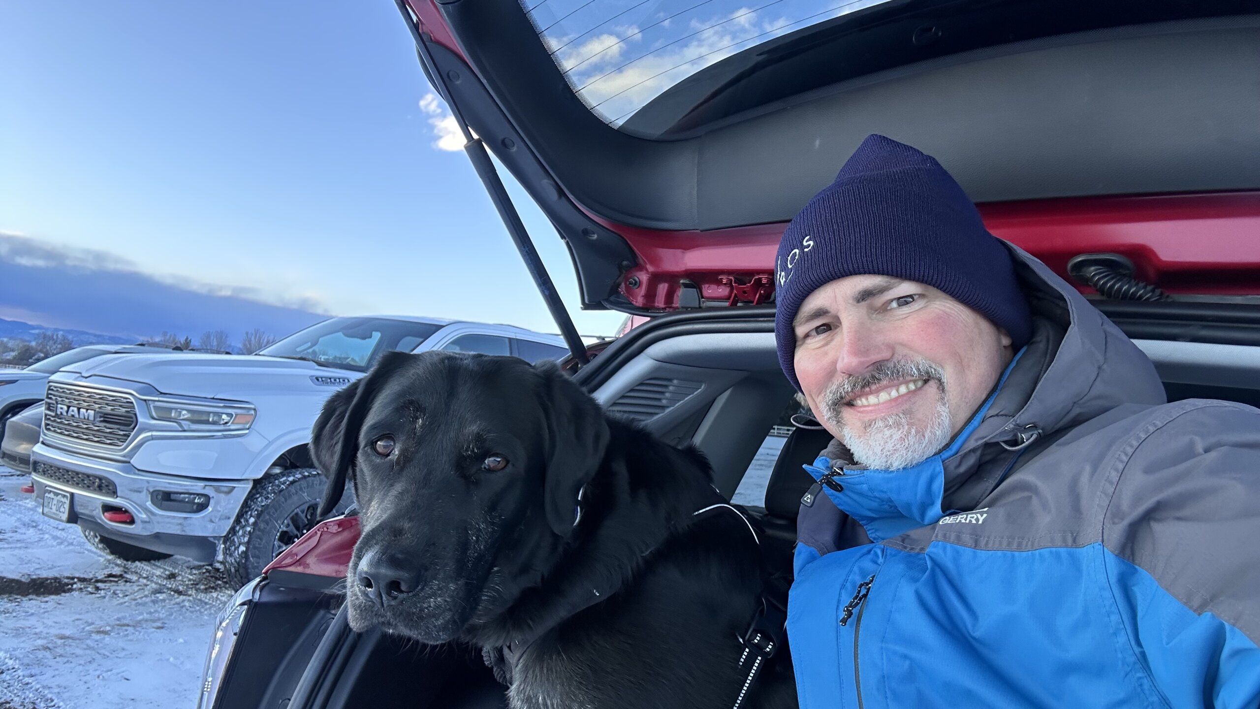 Man wearing a snow coat and hat sitting in the back of the car with a black and tan Labrador Retriever. Both look happy.