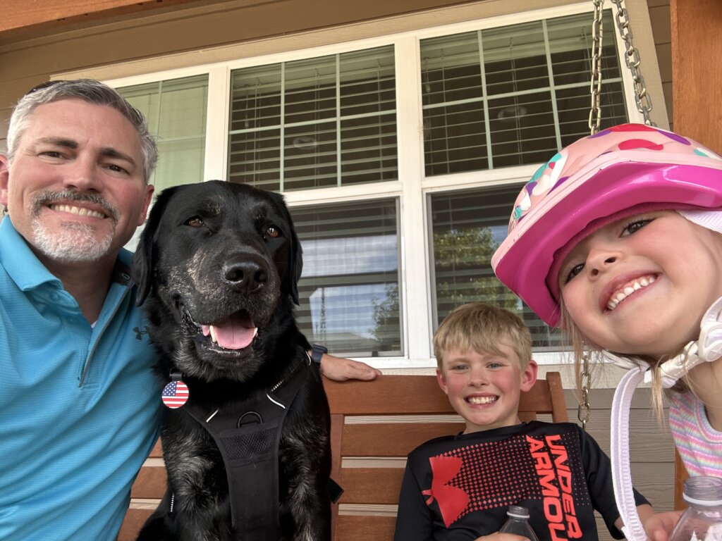 A man and two kids sitting with a black and tan Labrador Retriever Skilled Companion Dog.