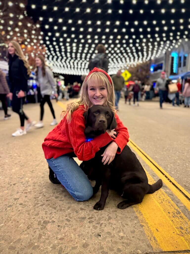 A Chocolate Labrador Retriever sitting with a young woman hugging her underneath bright lights.