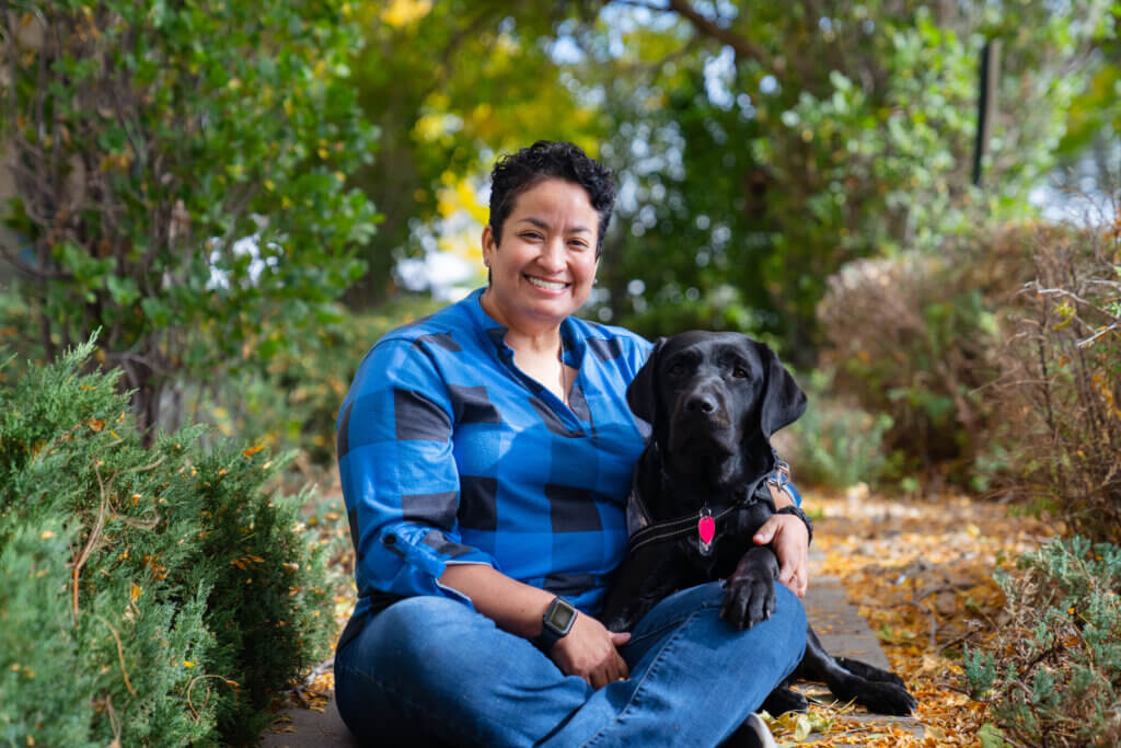 Female veteran wearing a blue plaid shirt sitting with her black Labrador service dog wearing a camo service dog vest.