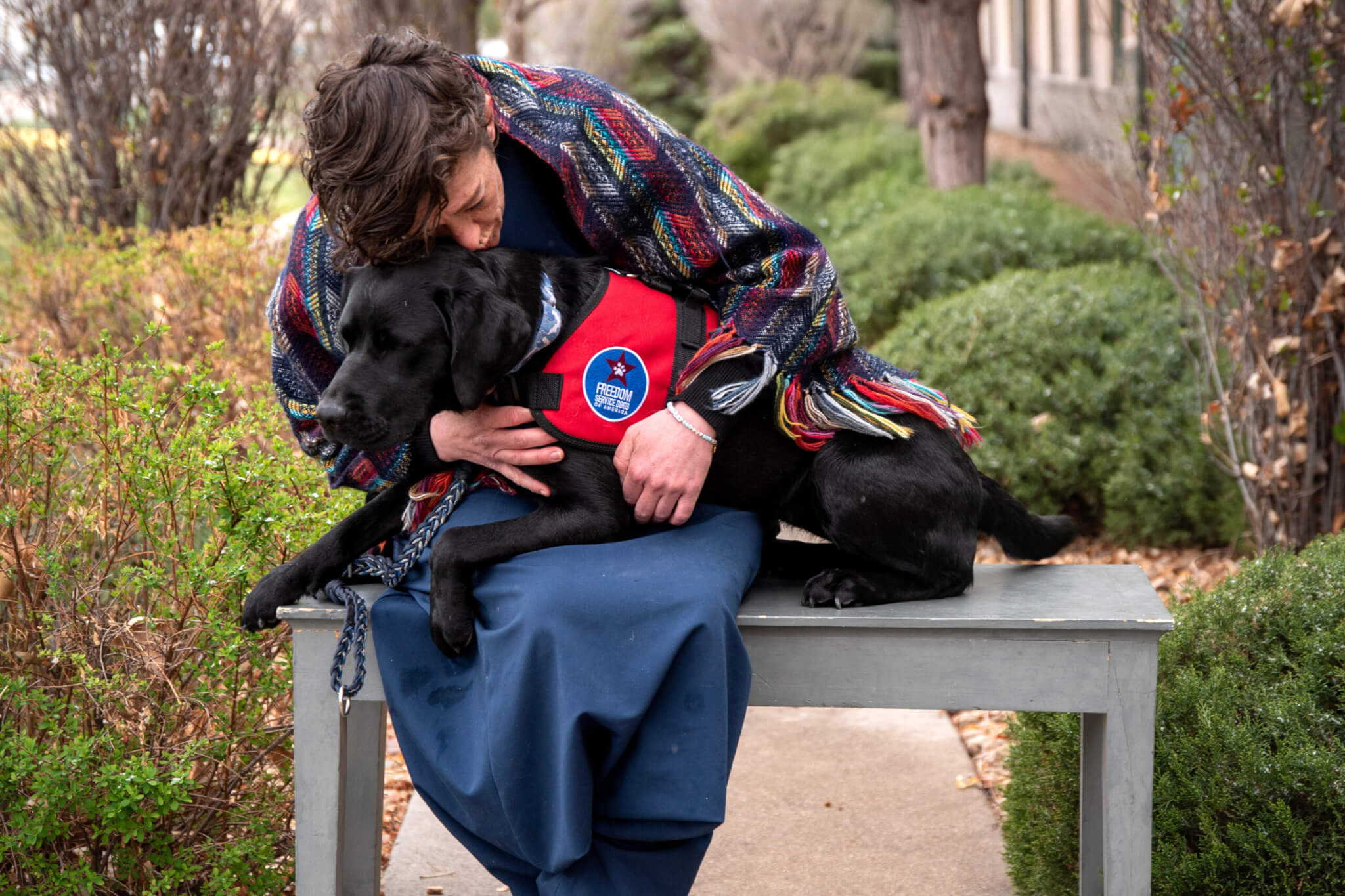 A woman leaning over to kiss a black Labrador Retriever wearing a Freedom Service Dogs service dog vest. The dog is performing deep pressure therapy.