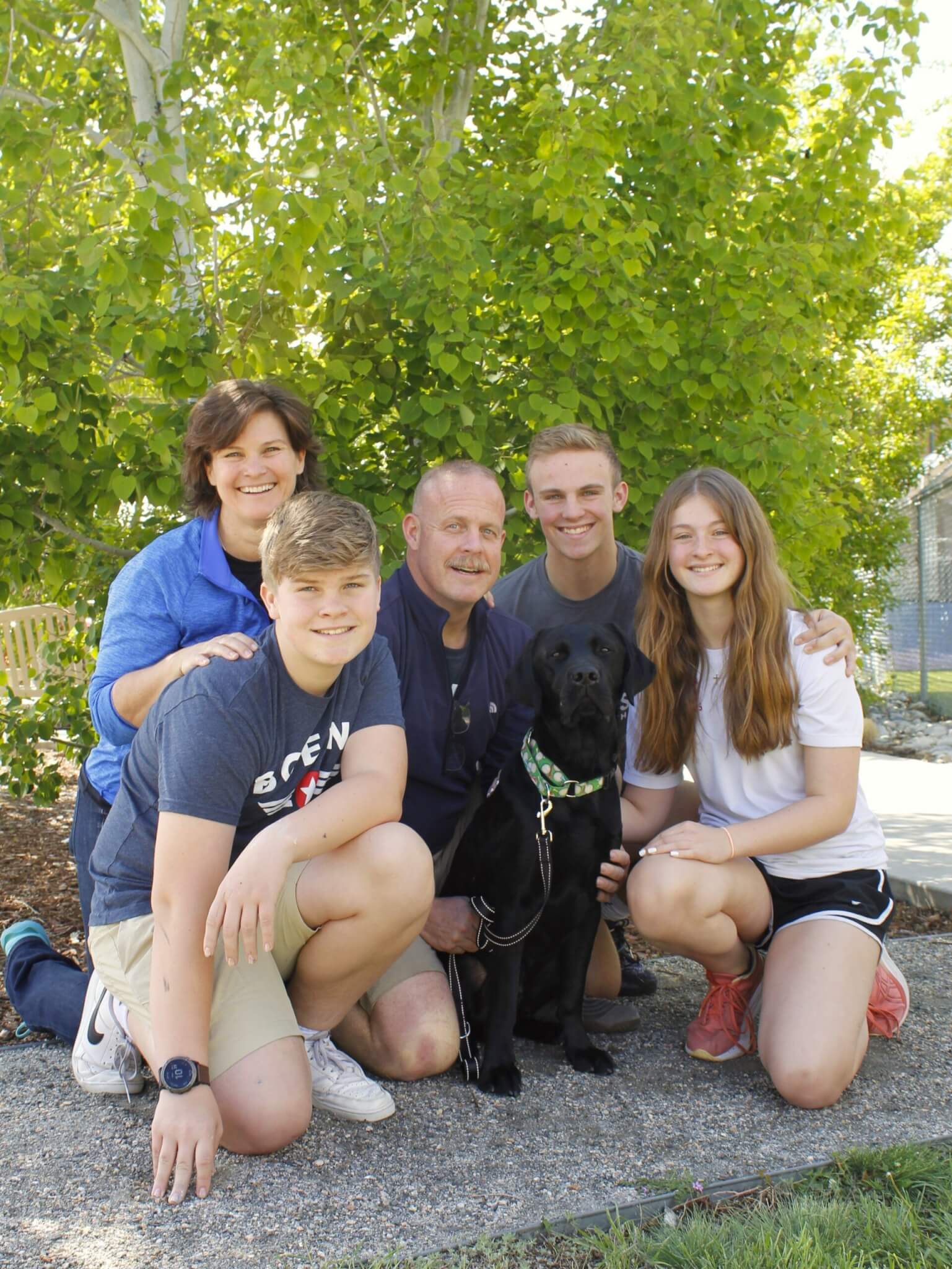 A Black Labrador Retriever sitting with a family that includes a woman, man, and three teenage children.