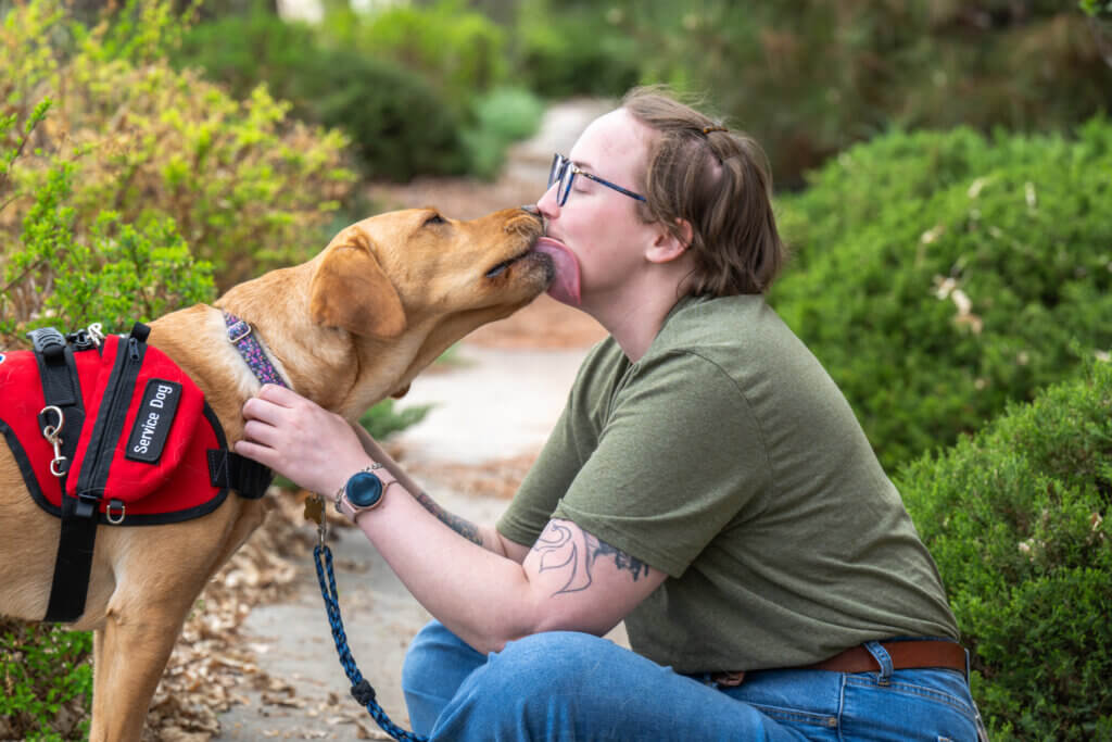A woman wearing a green shirt and glasses getting licked on the face by her yellow Labrador Retriever service dog.