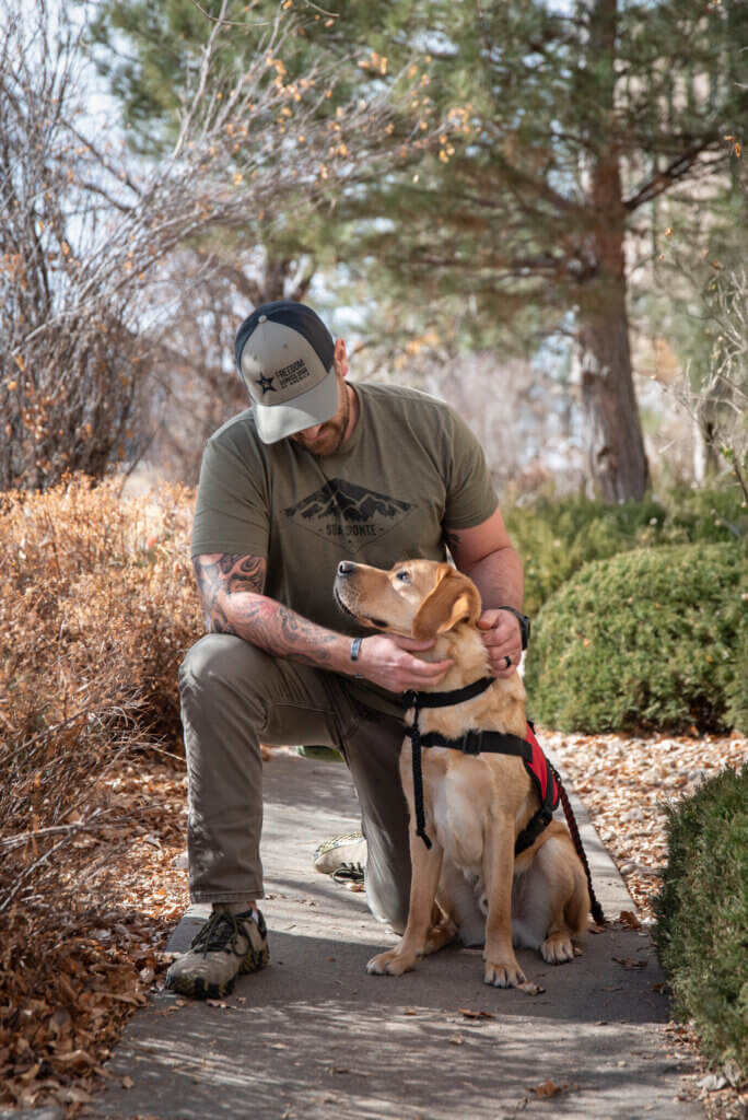 A man wearing a Freedom Service Dogs hat is kneeling on the ground and looking at his FSD Service Dog, who is a yellow Labrador Retriever wearing red service dog vest. The dog is looking at his person's eyes lovingly.