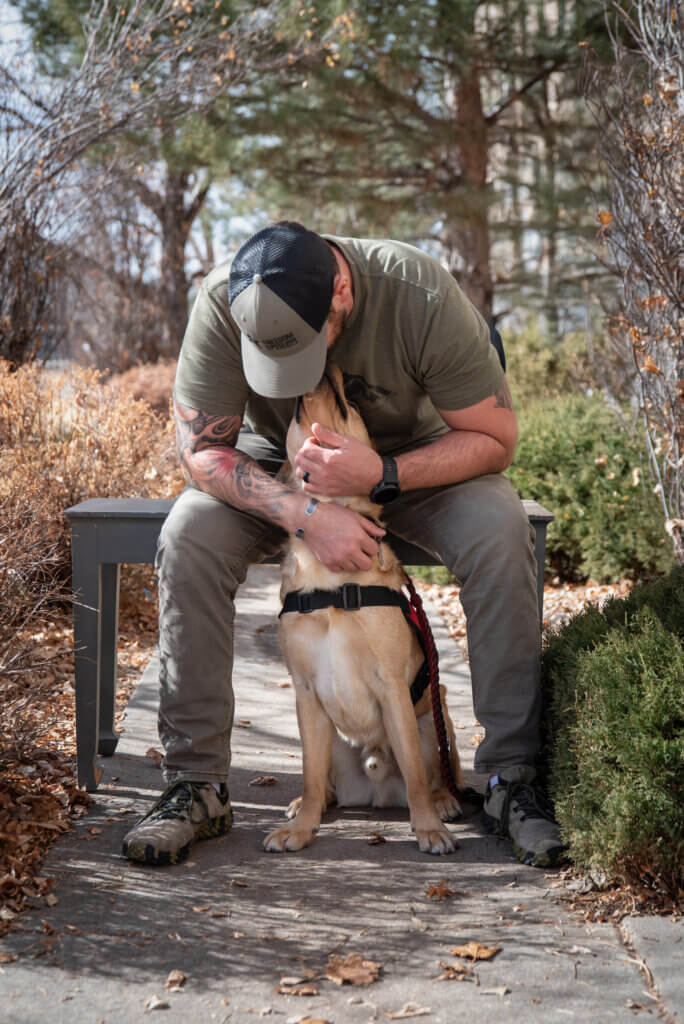 A U.S. Army veteran wearing a Freedom Service Dogs hat bending over to kiss his Freedom Service Dog, who is a yellow Labrador Retriever male wearing a red service dog vest.