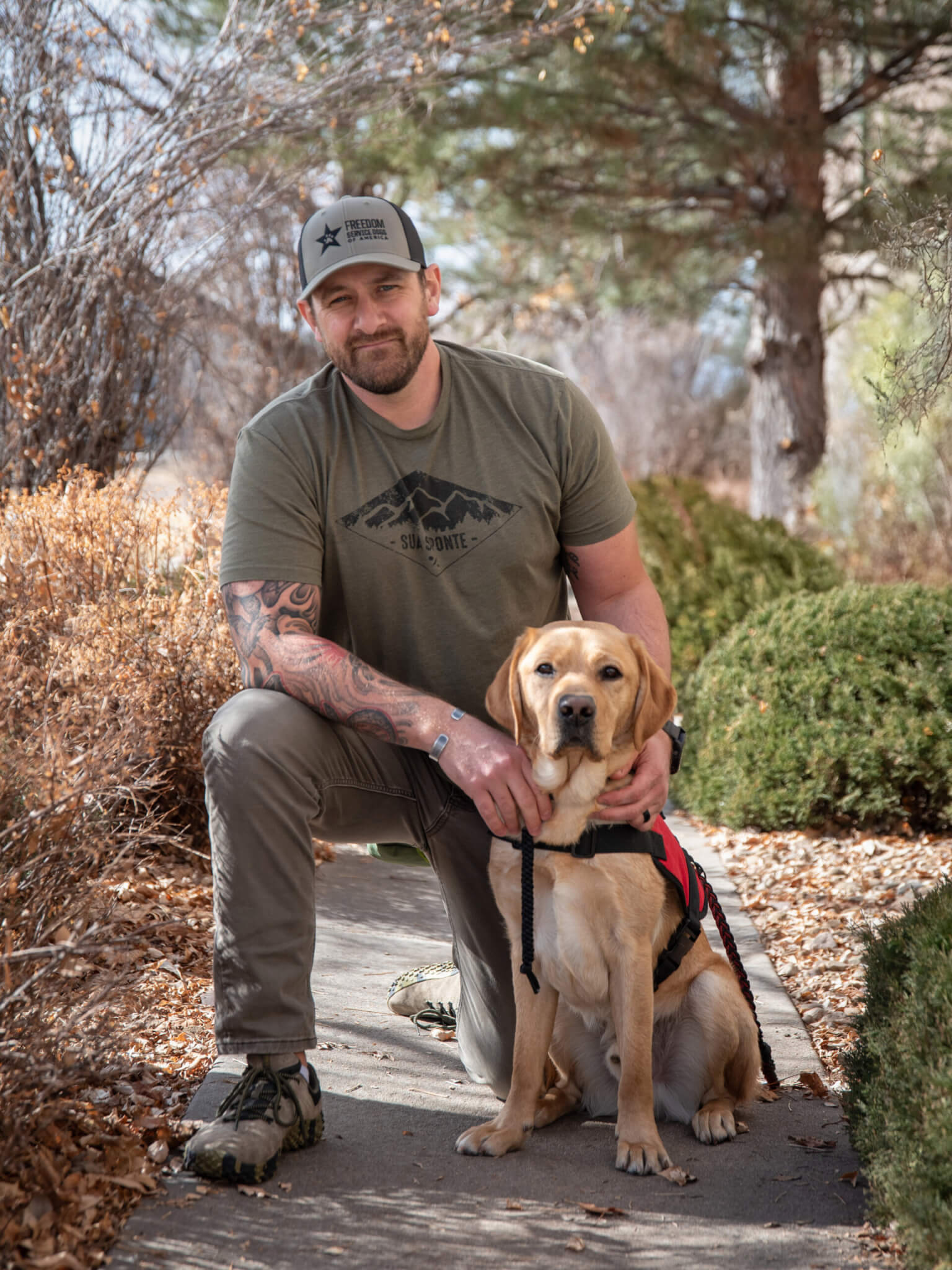 A U.S. Army veteran wearing a green shirt kneels beside his Freedom Service Dog- a yellow Labrador Retriever wearing a red service dog vest.