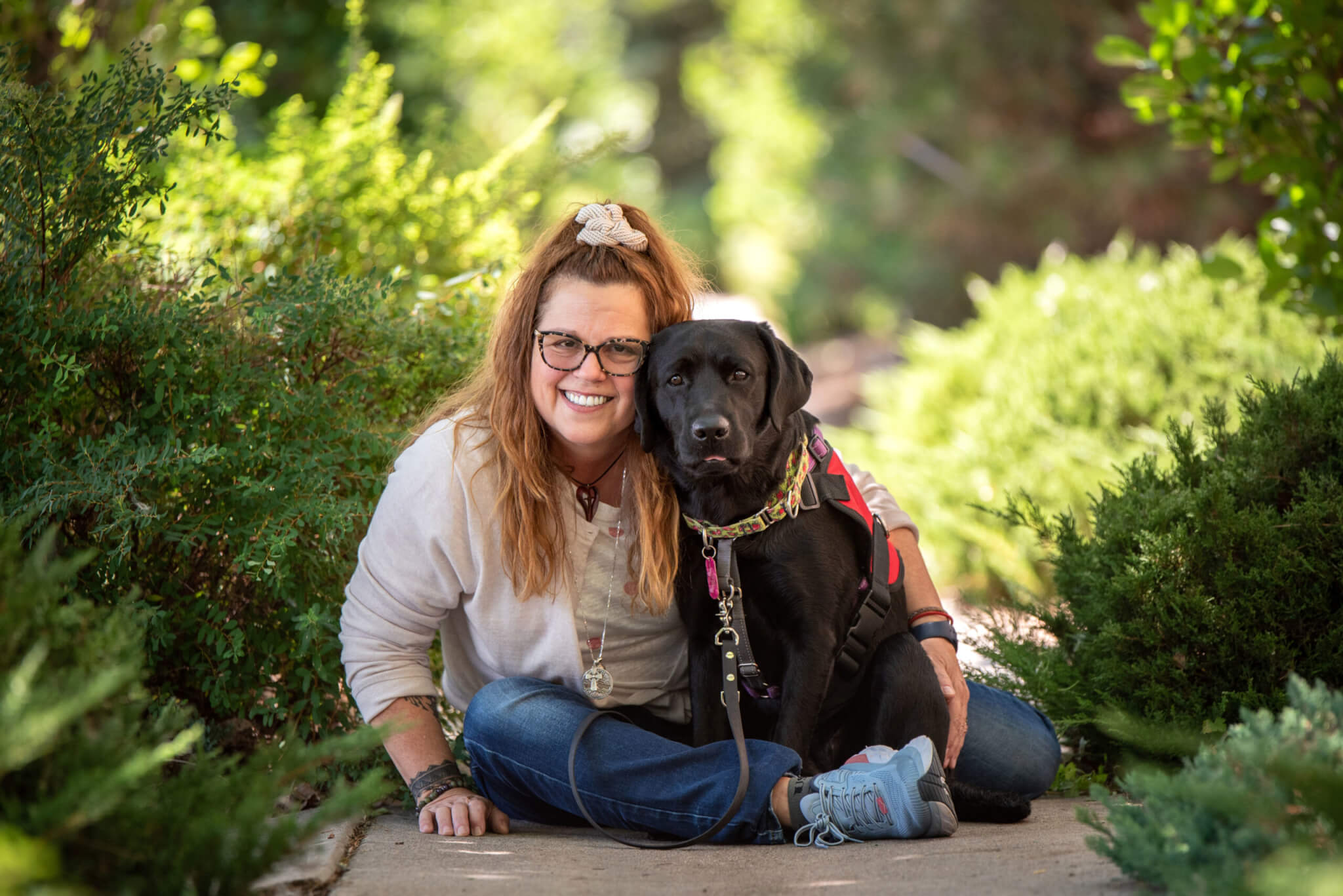 Lara, a veteran, sitting beside her black Labrador Retriever service dog, Cayenne, wearing a red FSD service dog vest.