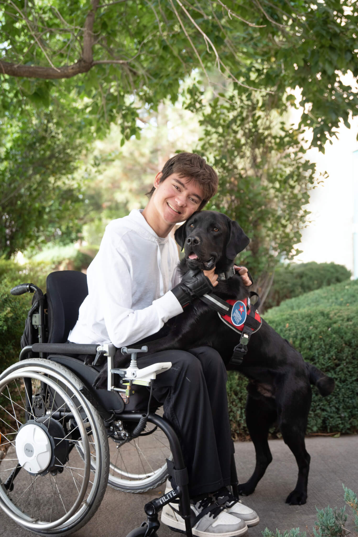 A young man in a wheelchair with a black Labrador Retriever service dog on his lap. 