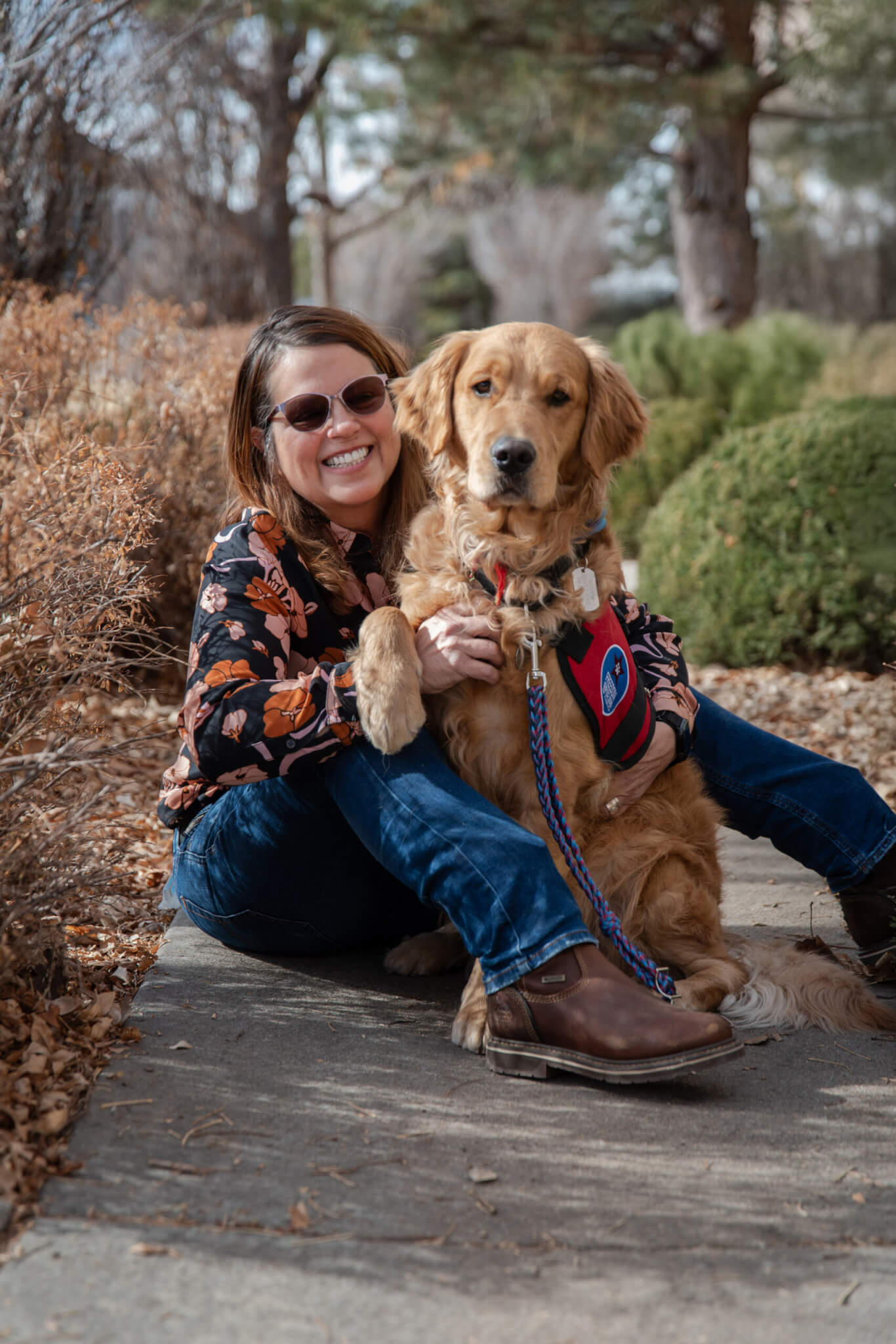 Lara sitting on a path outdoors with her FSD Service Dog Mushu, a golden retriever wearing a red Freedom Service Dogs service dog vest.