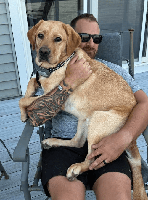 Nick wearing a gray t-shirt holding Dijon, a yellow Labrador Retriever, in his lap.