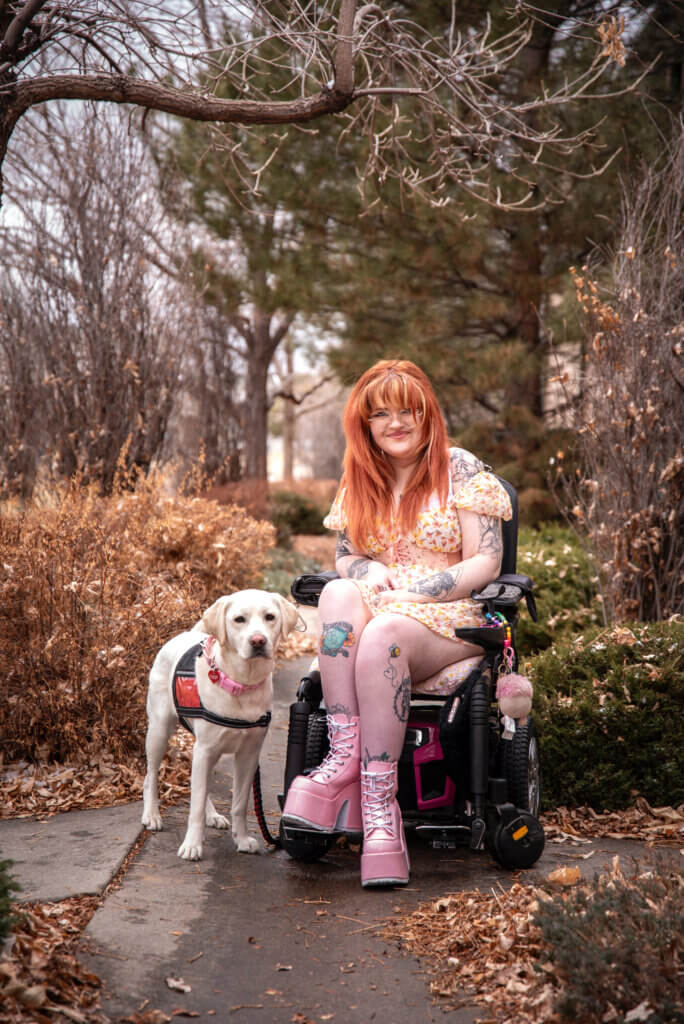 A young woman in a wheelchair named Kiya wearing a dress and pink boots. Beside her is her yellow Labrador retriever service dog, Cinema, wearing a red service dog vest from Freedom Service Dogs.