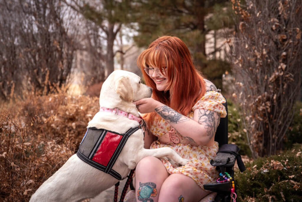 A young woman in a wheelchair named Kiya wears a white and yellow dress. She is smiling at her yellow Labrador retriever service dog, Cinema, who wears a red Freedom Service Dogs service dog vest.