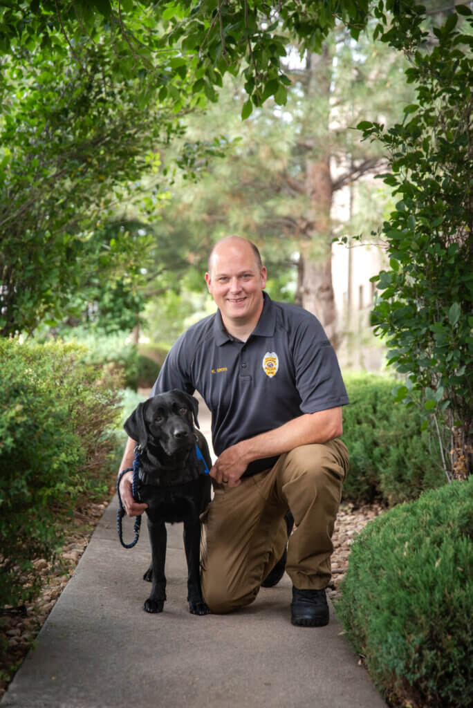 SRO Kevin in uniform standing beside his Black Labrador Retriever FSD Facility Dog, Jack. 