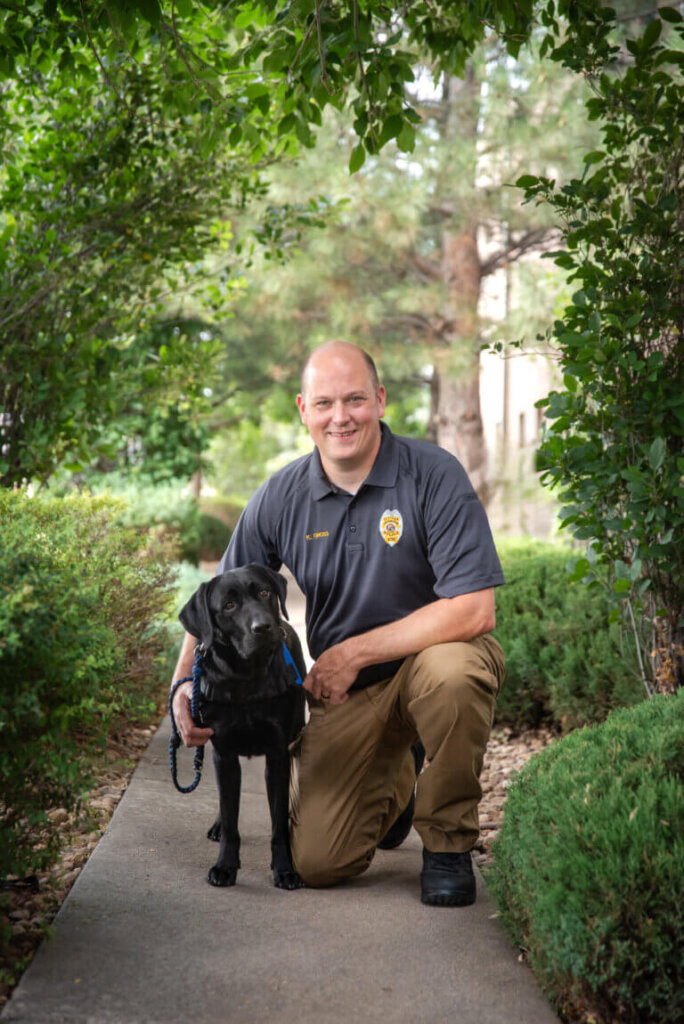 SRO Kevin in uniform standing beside his Black Labrador Retriever FSD Facility Dog, Jack.