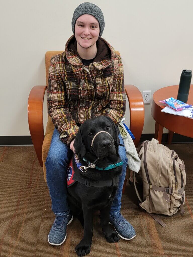 Vincent is sitting on a chair wearing a flannel, gray beanie, jeans, and tennis shoes. They are sitting with their service dog Rumble, who is wearing a red service dog vest.