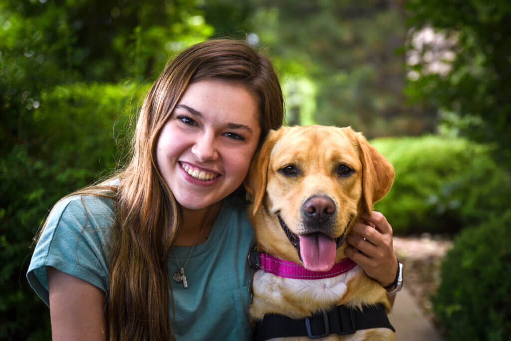 A young woman with light brown hair smiling and hugging a dog sitting beside her. The dog is a yellow Labrador retriever wearing a pink collar and red service dog vest.
