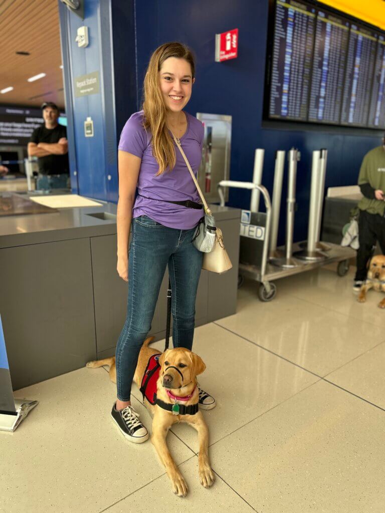 A young woman with light brown hair wearing a purple t-shirt, jeans, and black Converse standing at the airport. Between her feet is a yellow Labrador retriever wearing a red service dog vest.