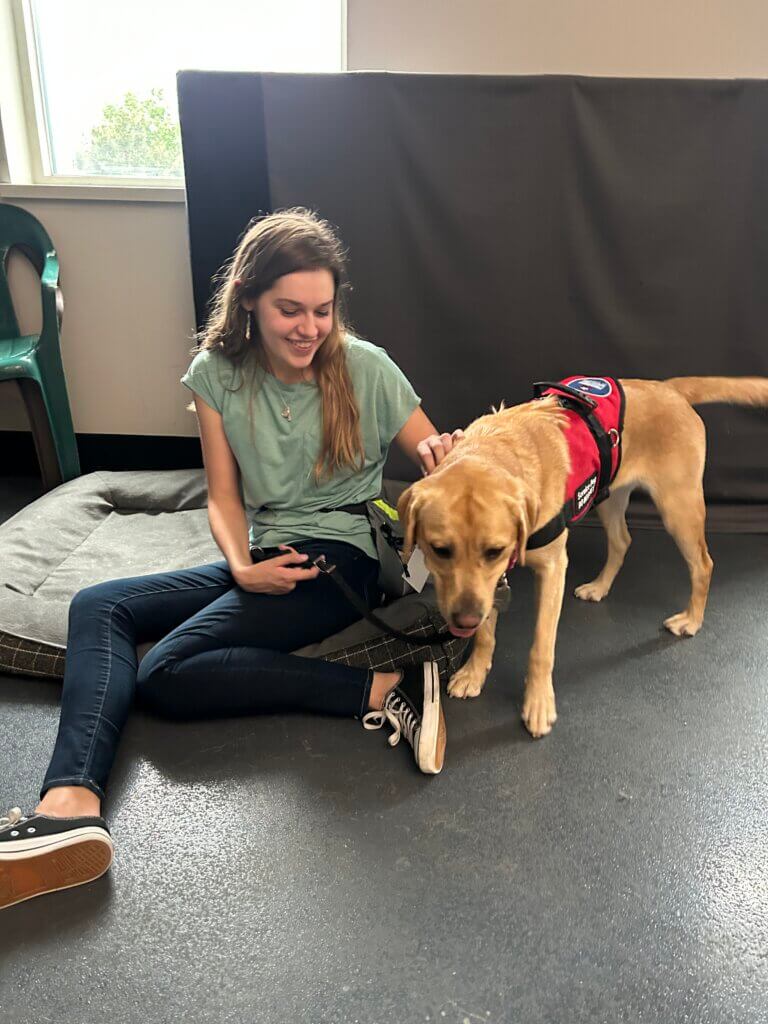 A young woman with light brown hair wearing jeans and a t-shirt sitting on the floor beside her yellow Labrador Retriever. The dog is standing and wearing a red service dog vest.