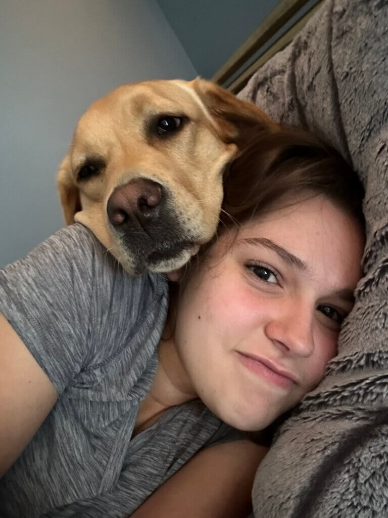 A young woman with light brown hair laying in bed. She is wearing a grey t-shirt. Resting her head on the young woman is a yellow Labrador retriever.