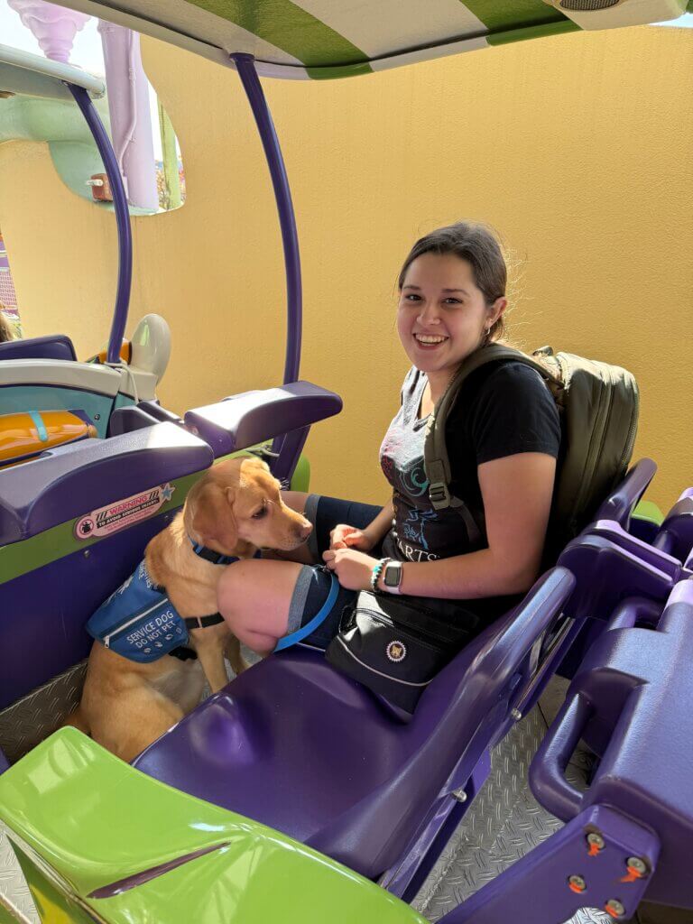 A young woman sitting on a purple cart. She is wearing a back back. Beside her is a yellow Labrador retriever wearing a service dog vest sitting at her feet.