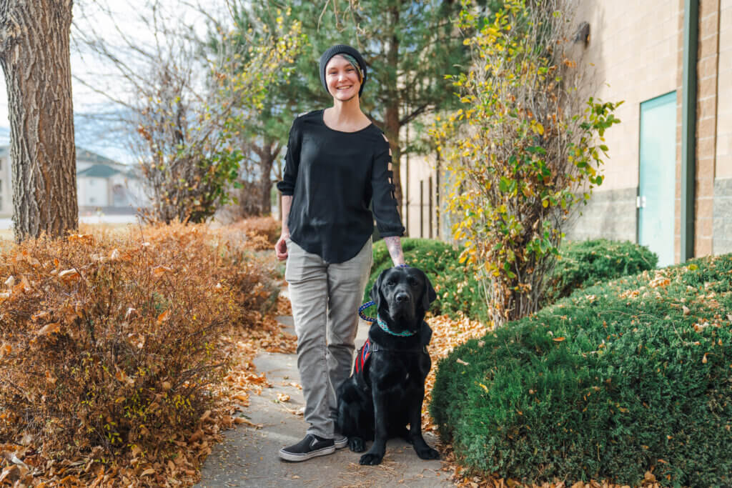 Vincent, an individual wearing a black shirt, cargo pants, black shoes, and a beanie, standing beside bushes and their black Labrador retriever. The dog is wearing a red service dog vest and a blue dog collar.