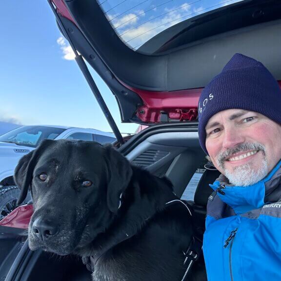 Man wearing a snow hat sitting in a car with a black and tan Labrador Retriever.