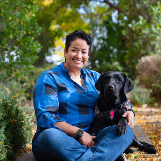 A woman sitting with her black Labrador Retriever service dog wearing a camo Freedom Service Dogs service dog vest.