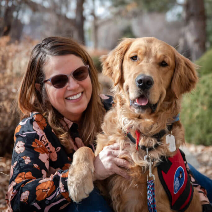 Woman wearing sunglasses with her golden retriever service dog Mushu wearing a red Freedom Service Dogs vest.