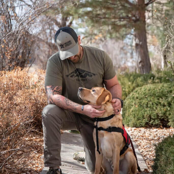 A man wearing a Freedom Service Dogs hat is kneeling on the ground and looking at his FSD Service Dog, who is a yellow Labrador Retriever wearing red service dog vest. The dog is looking at his person's eyes lovingly.